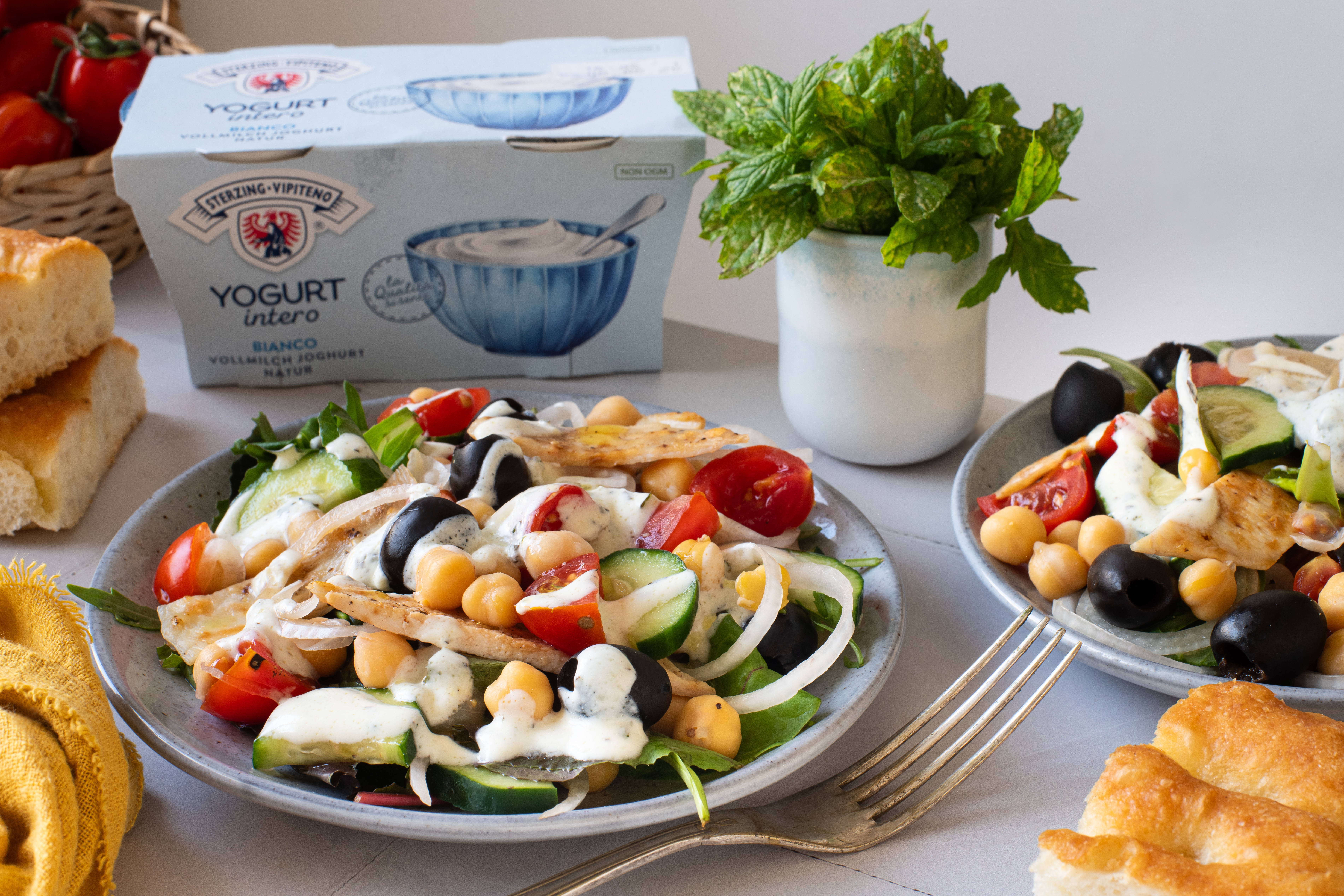 Zwei Teller mit Salat aus Hähnchenstreifen, Kichererbsen, Gurkenscheiben, Tomaten, schwarzen Oliven und Zwiebelringen mit Joghurtdressing, daneben Brot, ein Becher Minze und eine Packung Vollmilchjoghurt