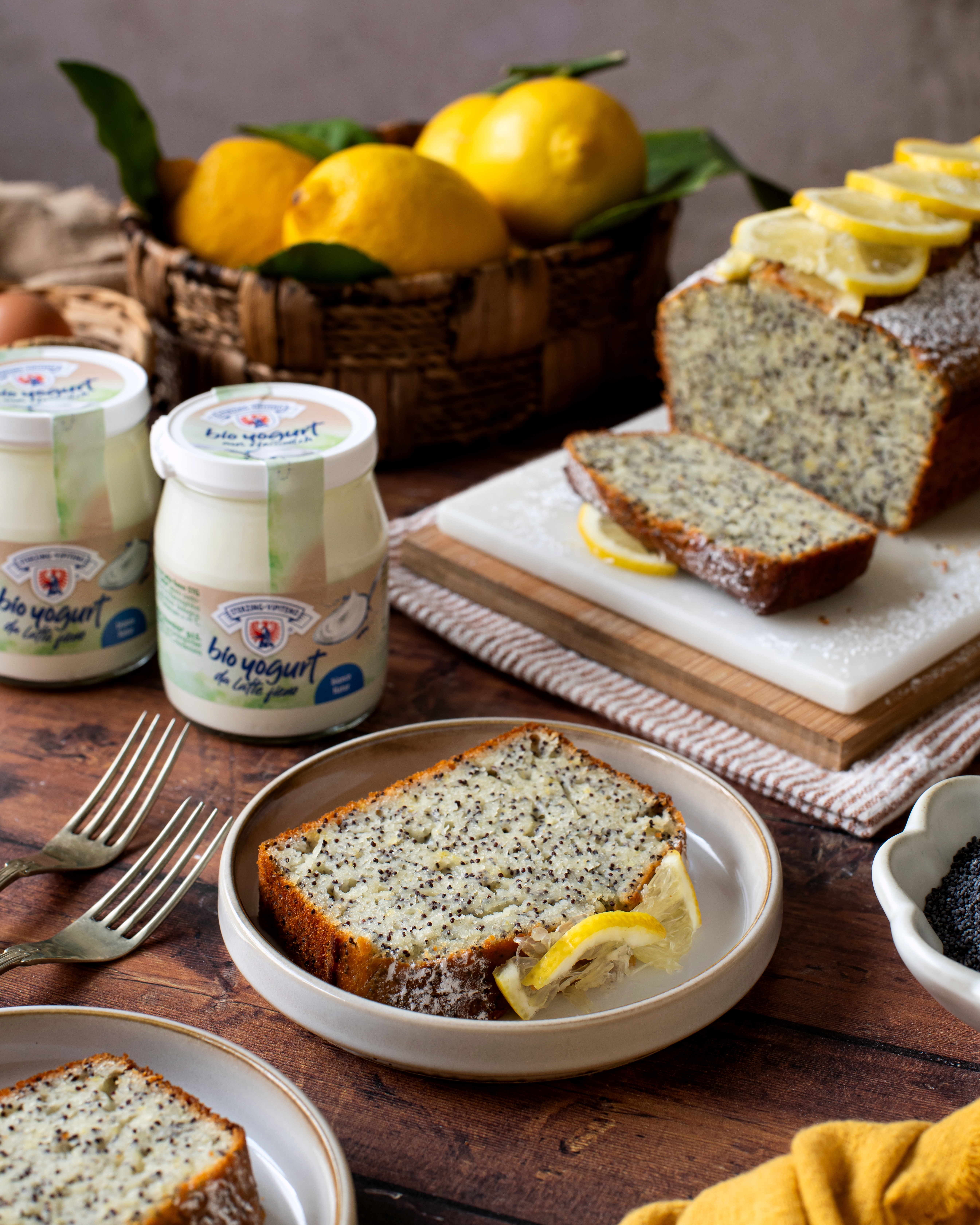 Mohn-Zitronenkuchen auf Teller mit Zitronenscheibe daneben, im Hintergrund ganzes Kuchenstück mit Zitronenscheiben, Joghurtgläser, Zitronen in Korb und Mohnsamen in Schale