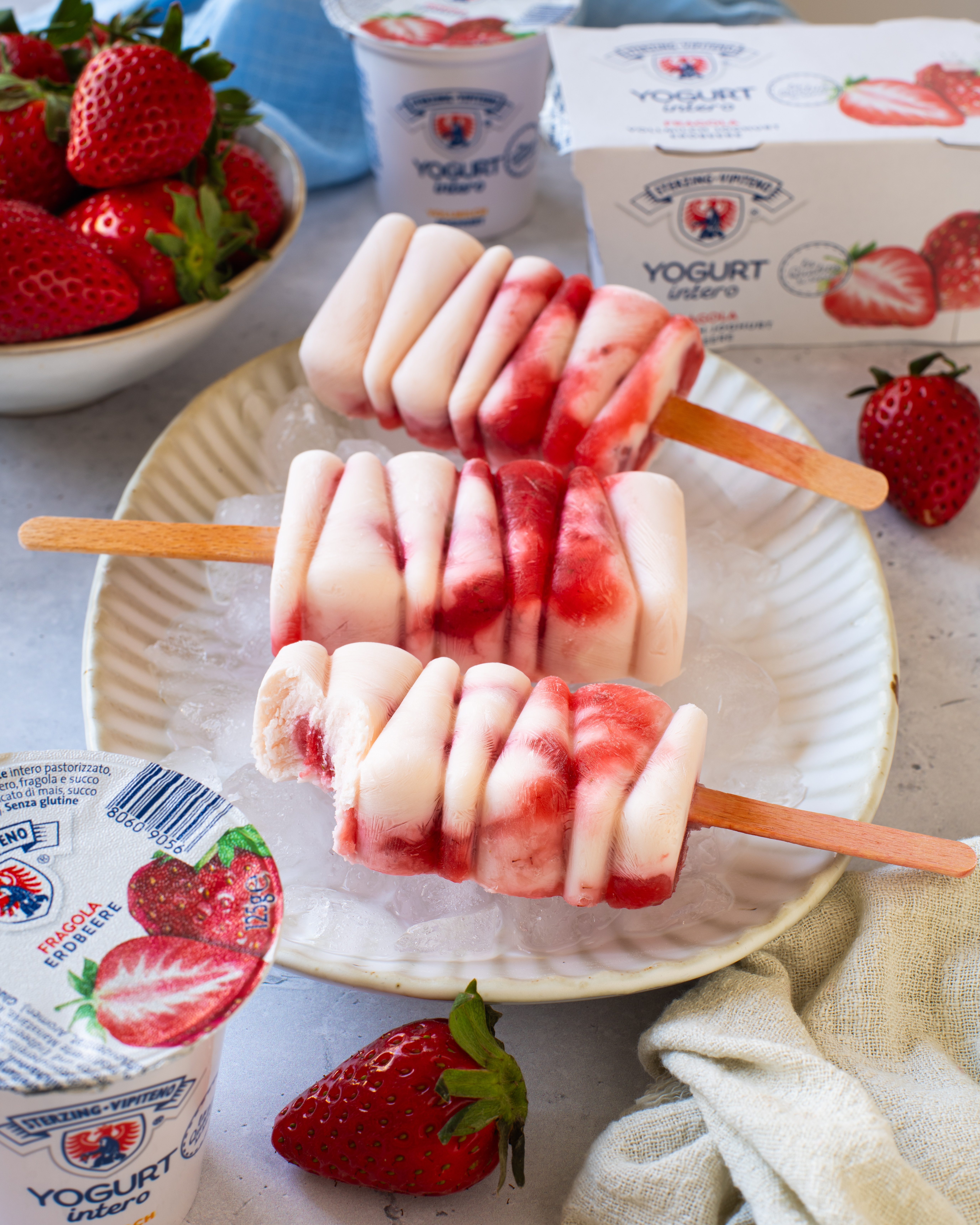 Drei Joghurt-Eis am Stiel mit Erdbeerstücken auf einem Teller mit Eiswürfeln, umgeben von Erdbeeren und Joghurtbechern