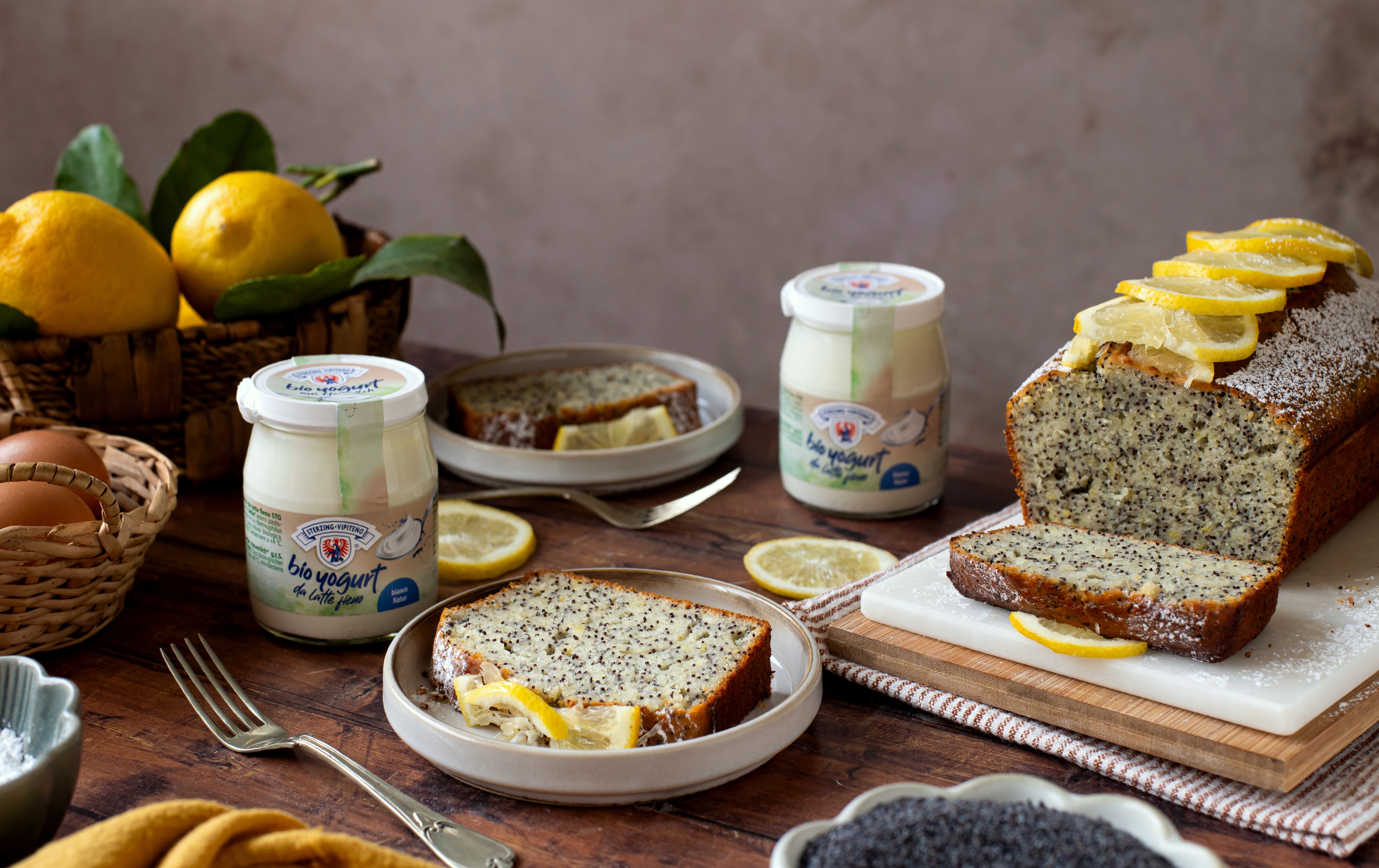 Mohn-Zitronenkuchen auf Teller mit Zitronenscheibe daneben, im Hintergrund ganzes Kuchenstück mit Zitronenscheiben, Joghurtgläser, Zitronen in Korb und Mohnsamen in Schale