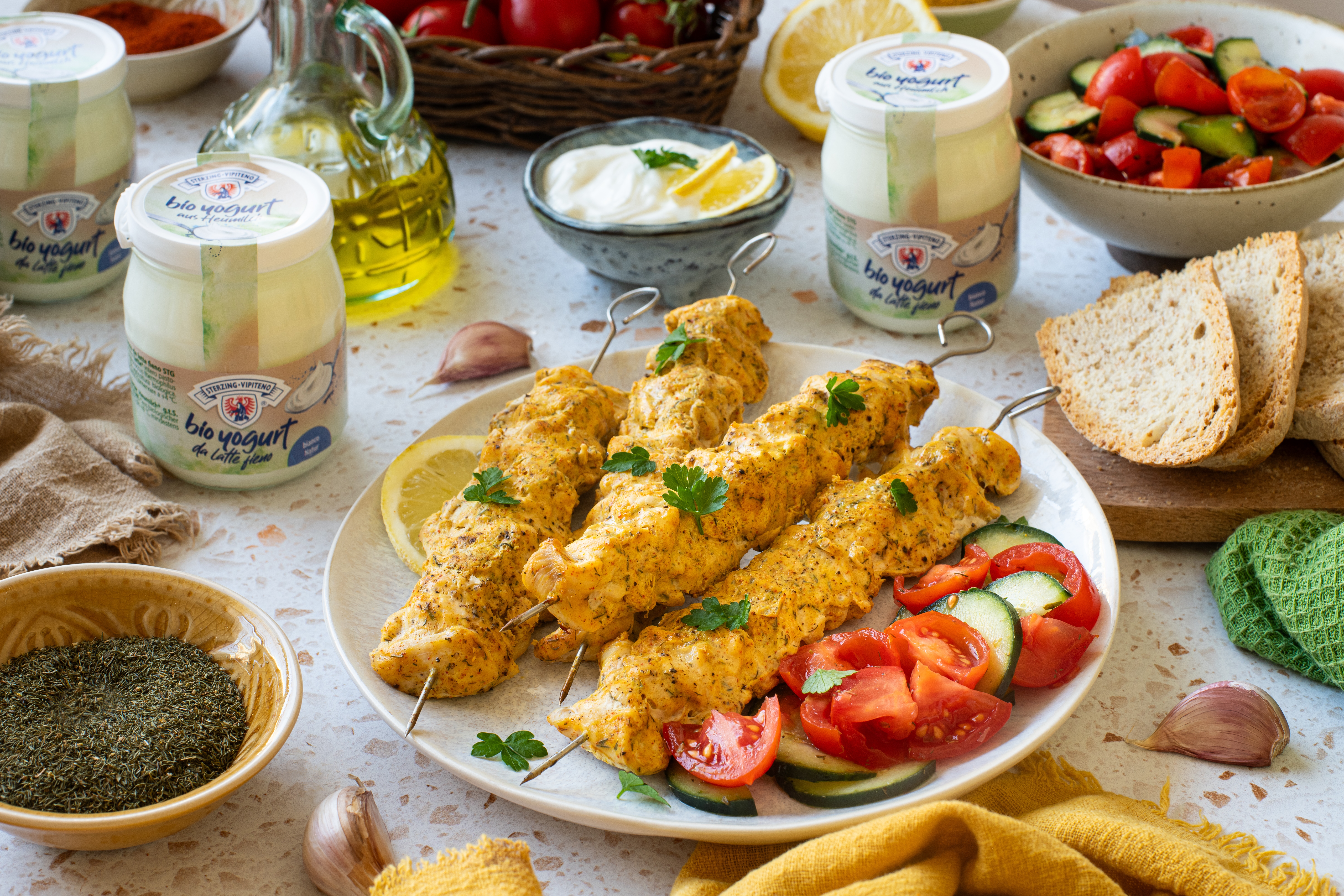 Teller mit vier Hähnchenspießen, garniert mit frischer Petersilie, dazu Tomaten- und Gurkenscheiben, umgeben von Joghurtgläsern, Knoblauchzehen, Olivenöl, Brot und einem Salat in Schalen
