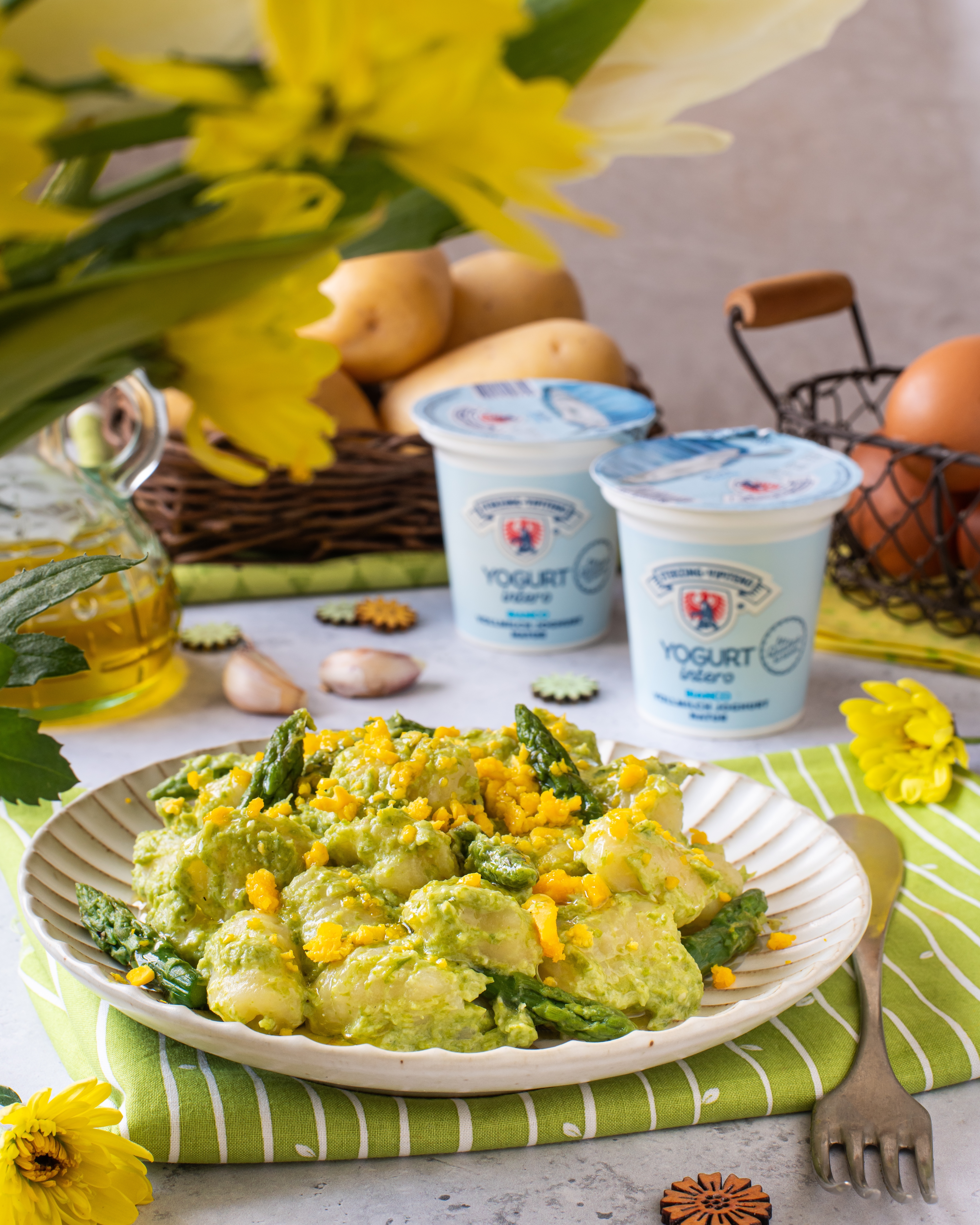 Teller mit Gnocchi in grüner Sauce und Mais, umgeben von Kartoffeln im Korb, Joghurtbechern, Salz- und Pfefferstreuern, Olivenölflasche, Knoblauchzehe, Eiern im Drahtkorb und gelben Blumen auf Tisch mit grün-weiß gestreifter Serviette
