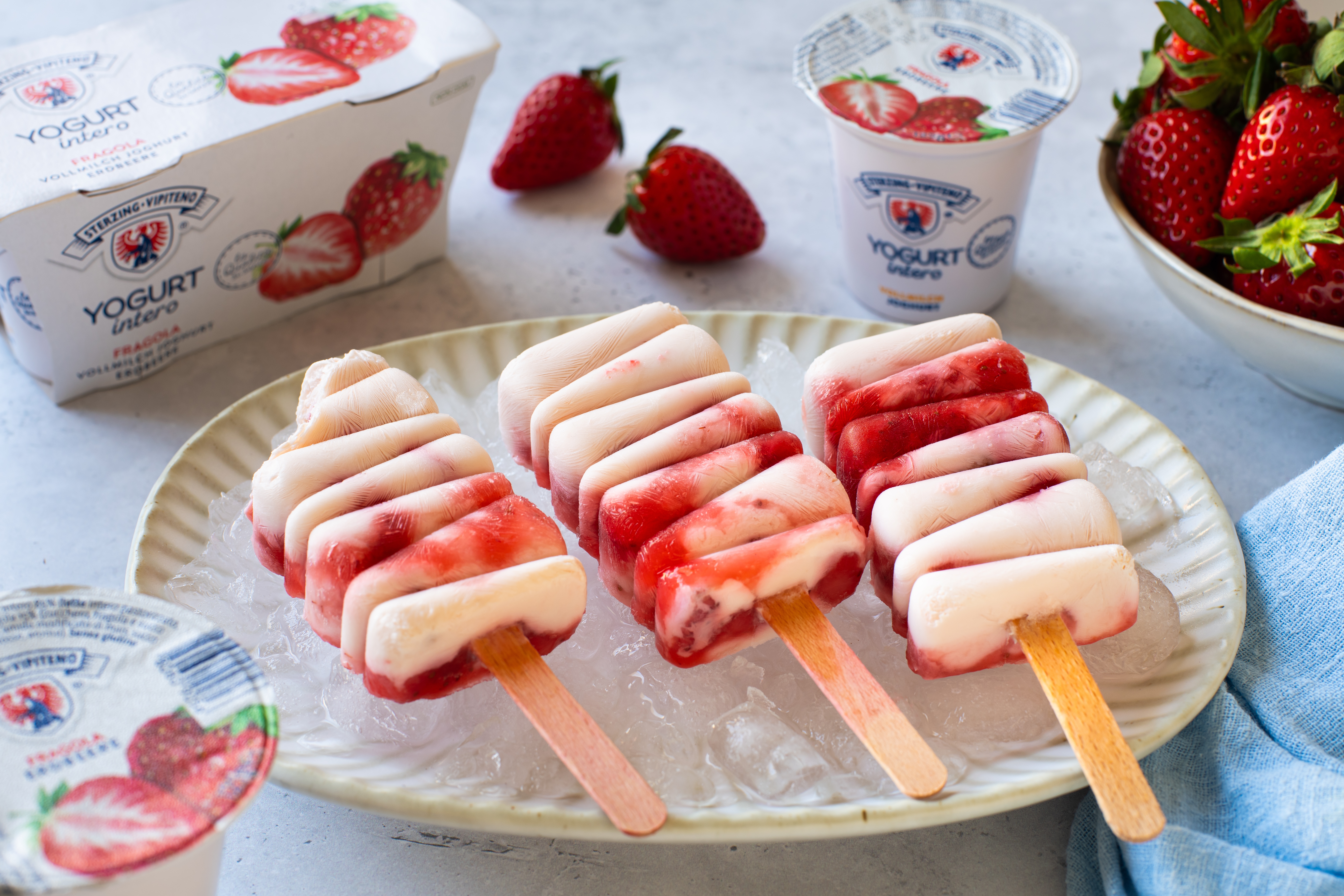 Drei Joghurt-Eis am Stiel mit Erdbeerstücken auf einem Teller mit Eiswürfeln, umgeben von Erdbeeren und Joghurtbechern
