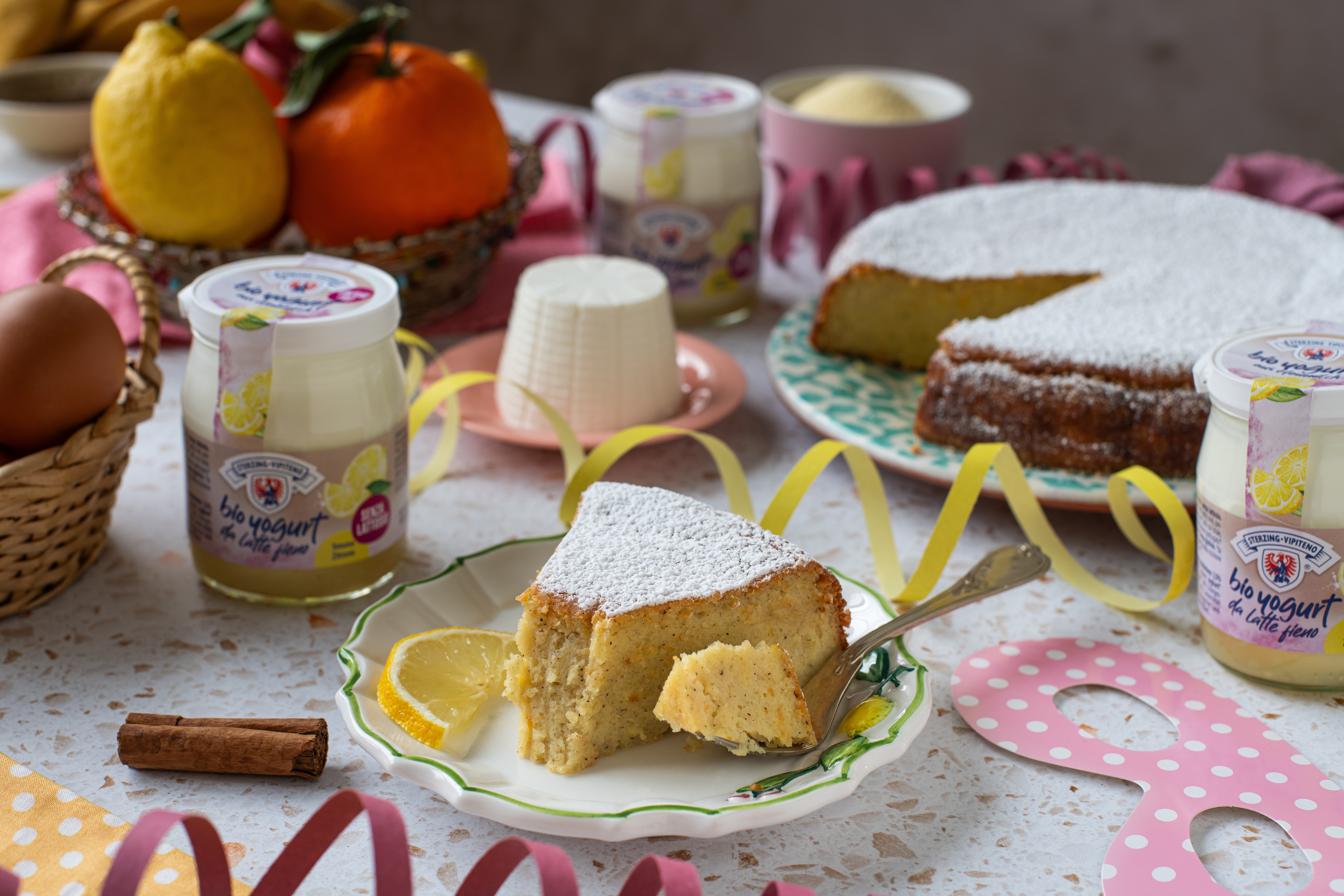 Stück mit Puderzucker bestäubter Kuchen auf Teller mit Zitronenscheiben und Gabel daneben Joghurtgläser, Eierkorb, Zimtstange und Obstkorb auf Tisch mit Tischdecke und Deko