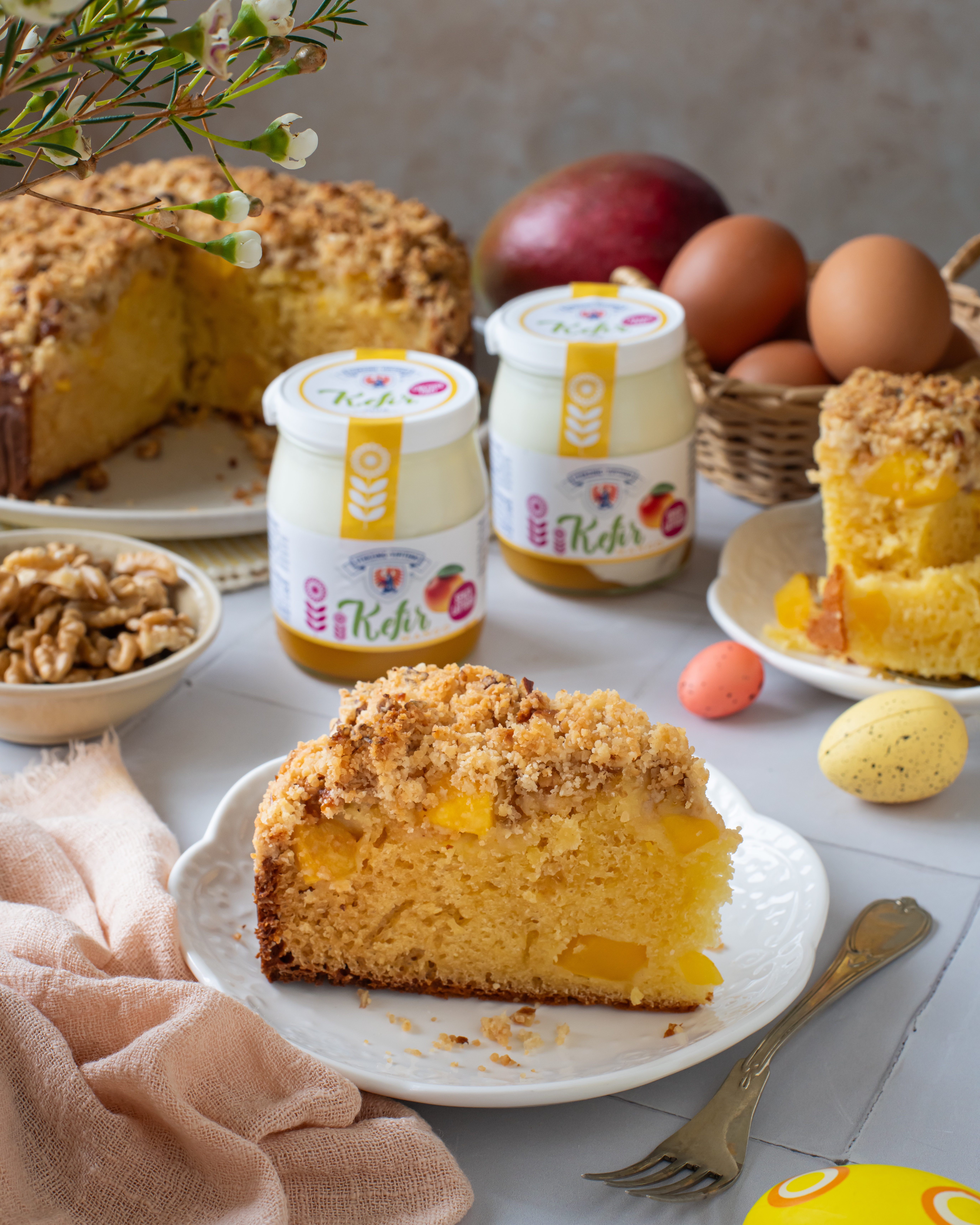 Stück Mango-Crumble-Kuchen auf weißem Teller mit Gabel umgeben von Kefir-Gläsern, Eiern, Walnüssen, Mango und Blumen