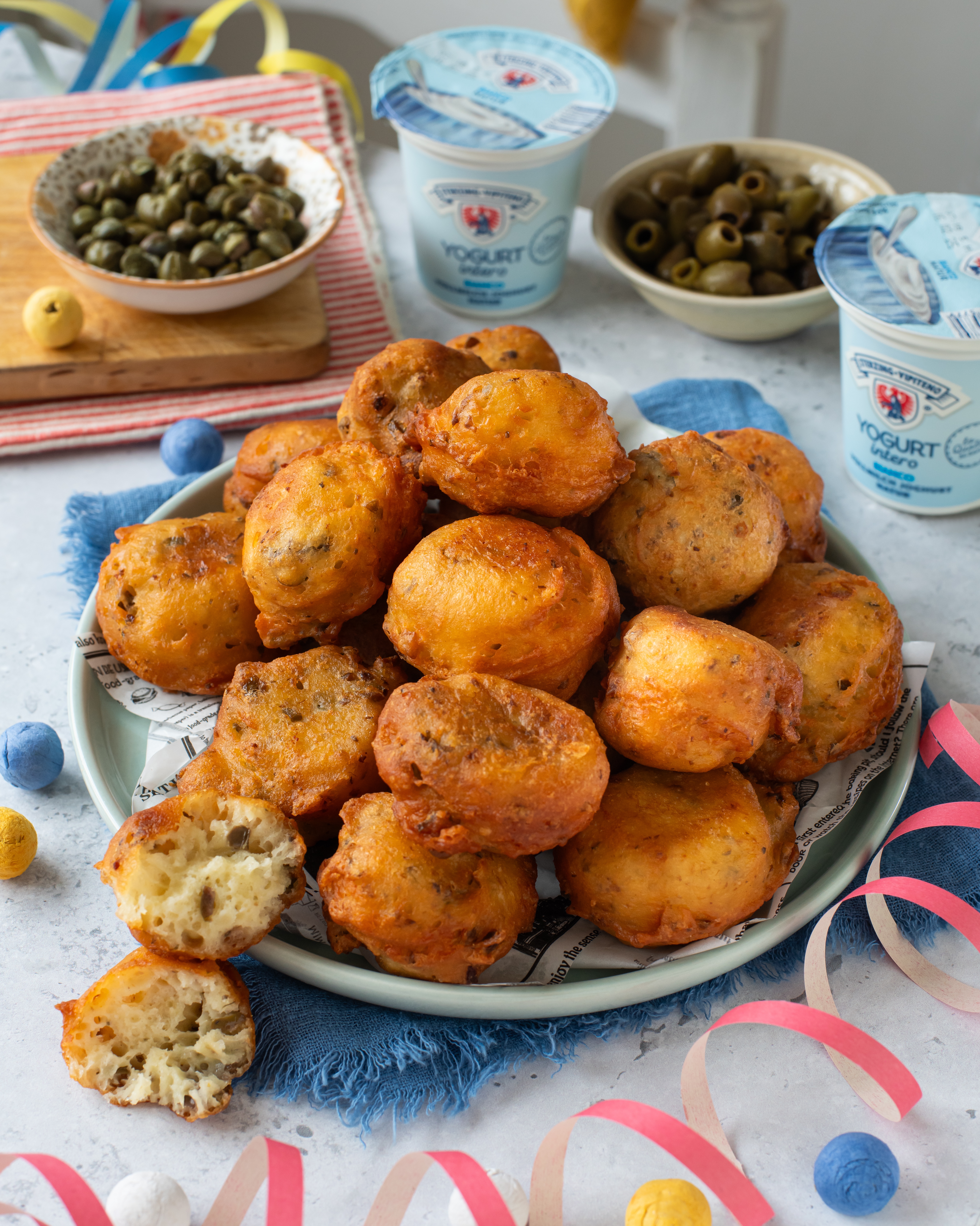 Teller mit frittierten Teigbällchen auf blauem Stoff, umgeben von Joghurtbechern, Oliven, Kapern, Eiern und bunten Papierstreifen