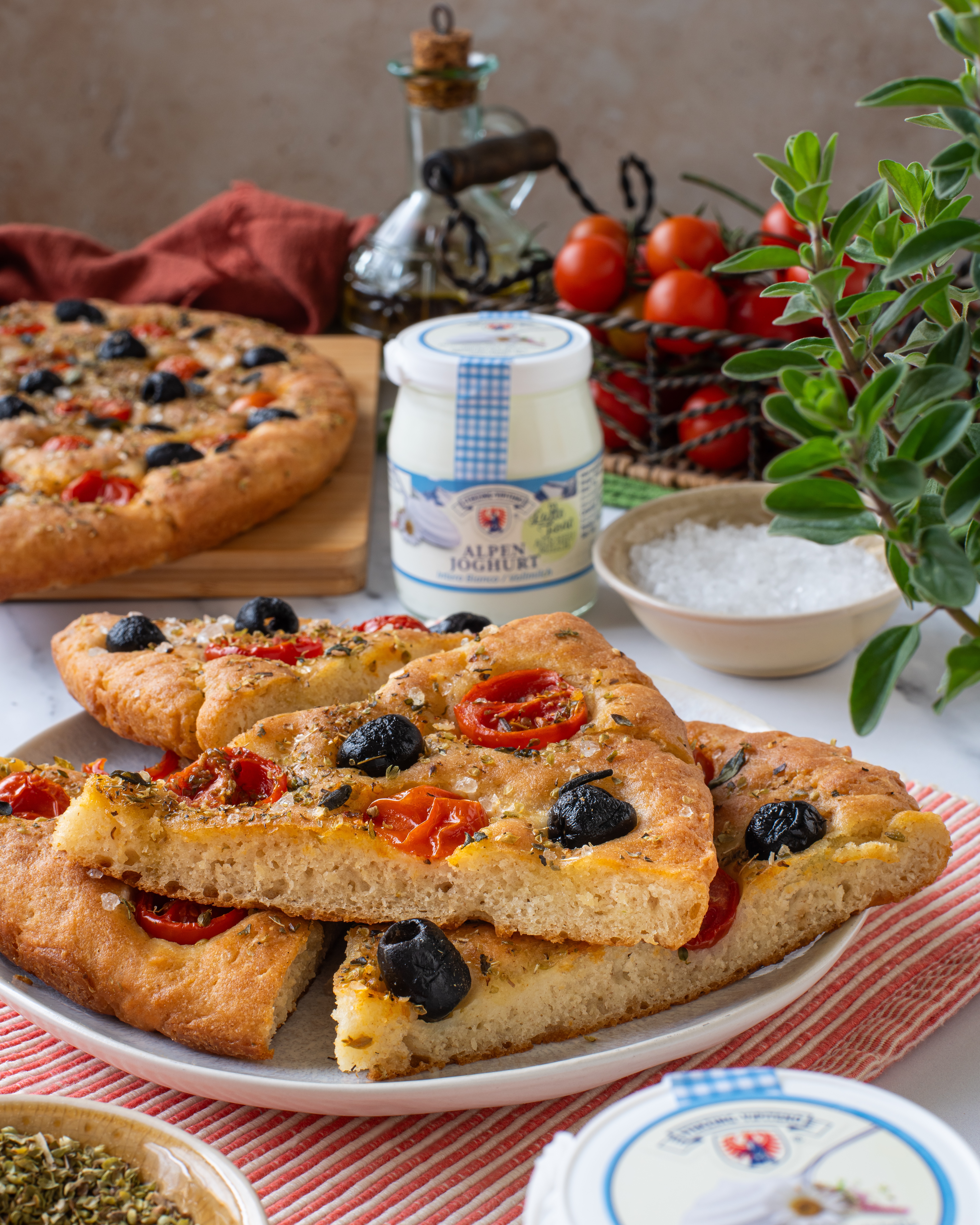 Dreieckige Stücke Focaccia mit Tomaten und schwarzen Oliven auf einem weißen Teller, umgeben von Joghurtgläsern und einem Korb mit Kirschtomaten