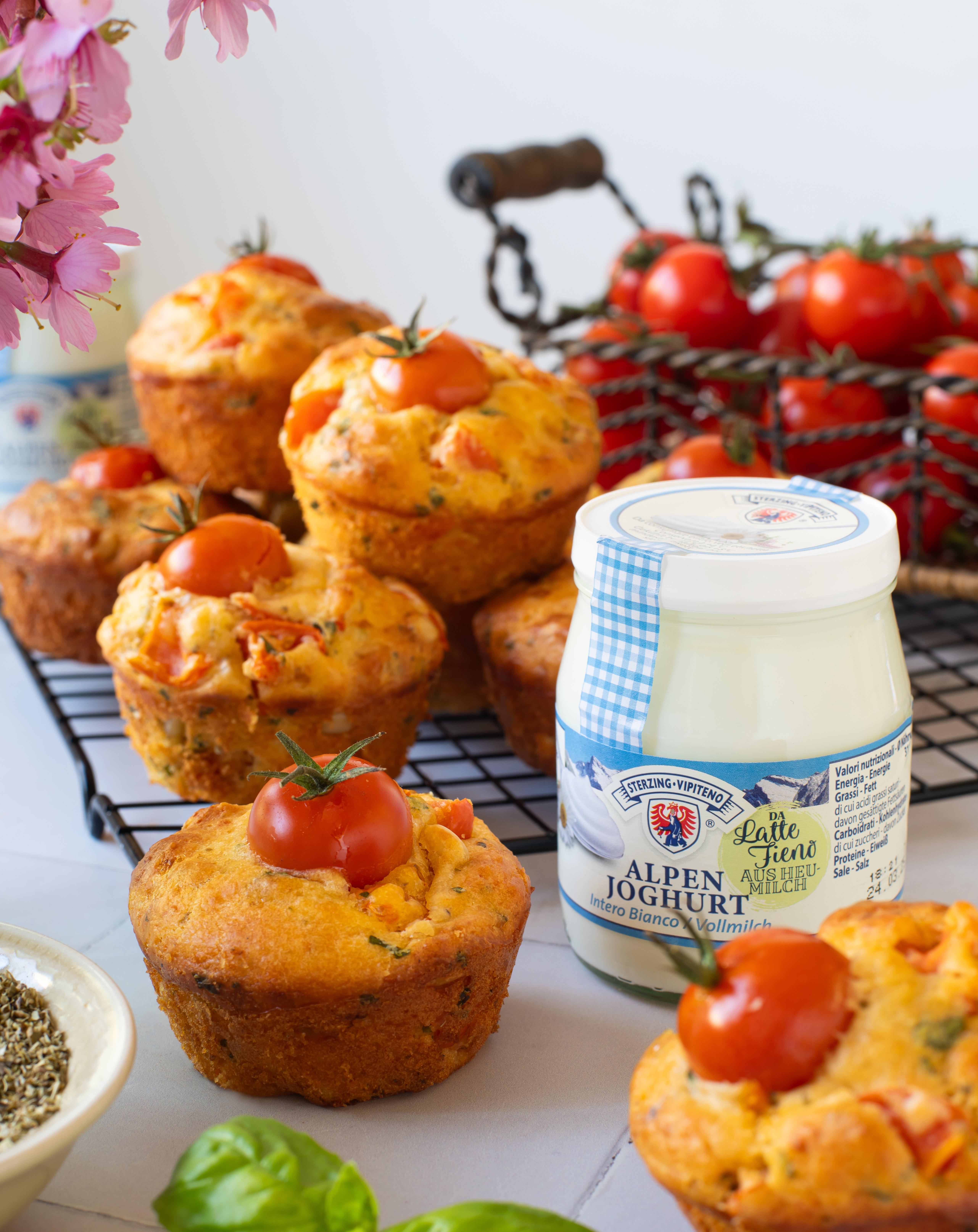 Mehrere herzhafte Muffins mit Kirschtomaten auf einem schwarzen Abkühlgitter, daneben ein Glas Alpenjoghurt und ein Korb mit Eiern