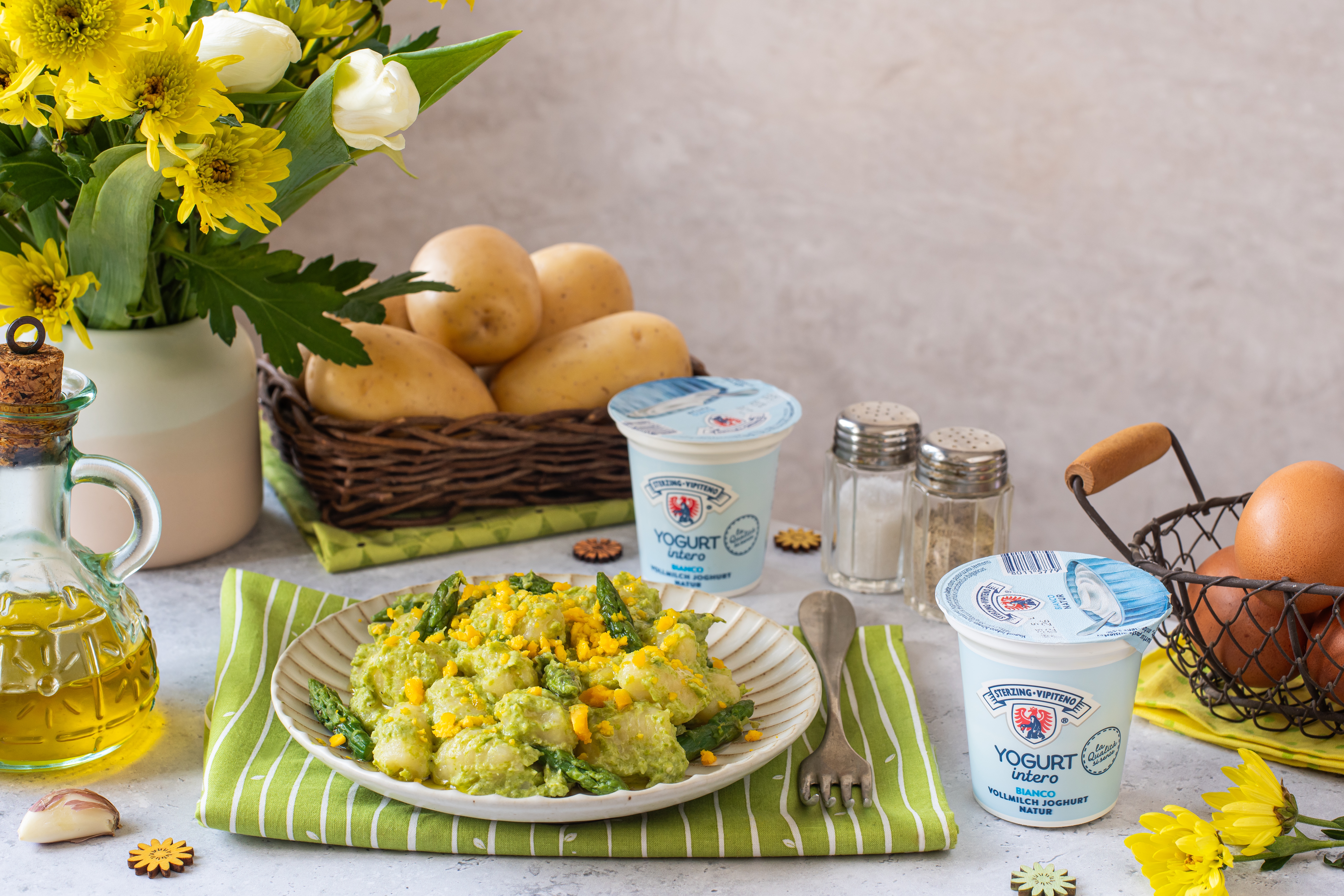 Teller mit Gnocchi in grüner Sauce und Mais, umgeben von Kartoffeln im Korb, Joghurtbechern, Salz- und Pfefferstreuern, Olivenölflasche, Knoblauchzehe, Eiern im Drahtkorb und gelben Blumen auf Tisch mit grün-weiß gestreifter Serviette