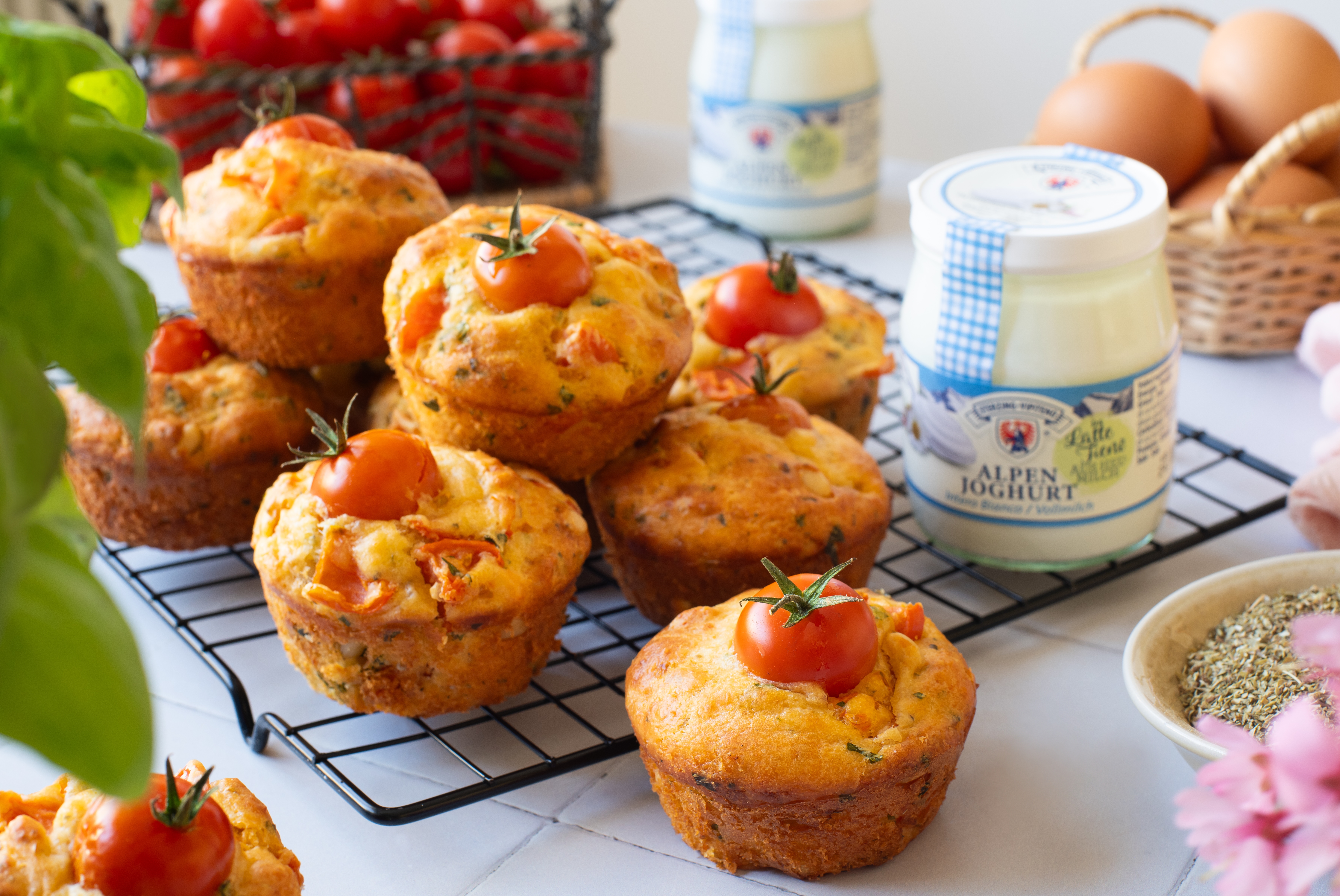 Mehrere herzhafte Muffins mit Kirschtomaten auf einem schwarzen Abkühlgitter, daneben ein Glas Alpenjoghurt und ein Korb mit Eiern
