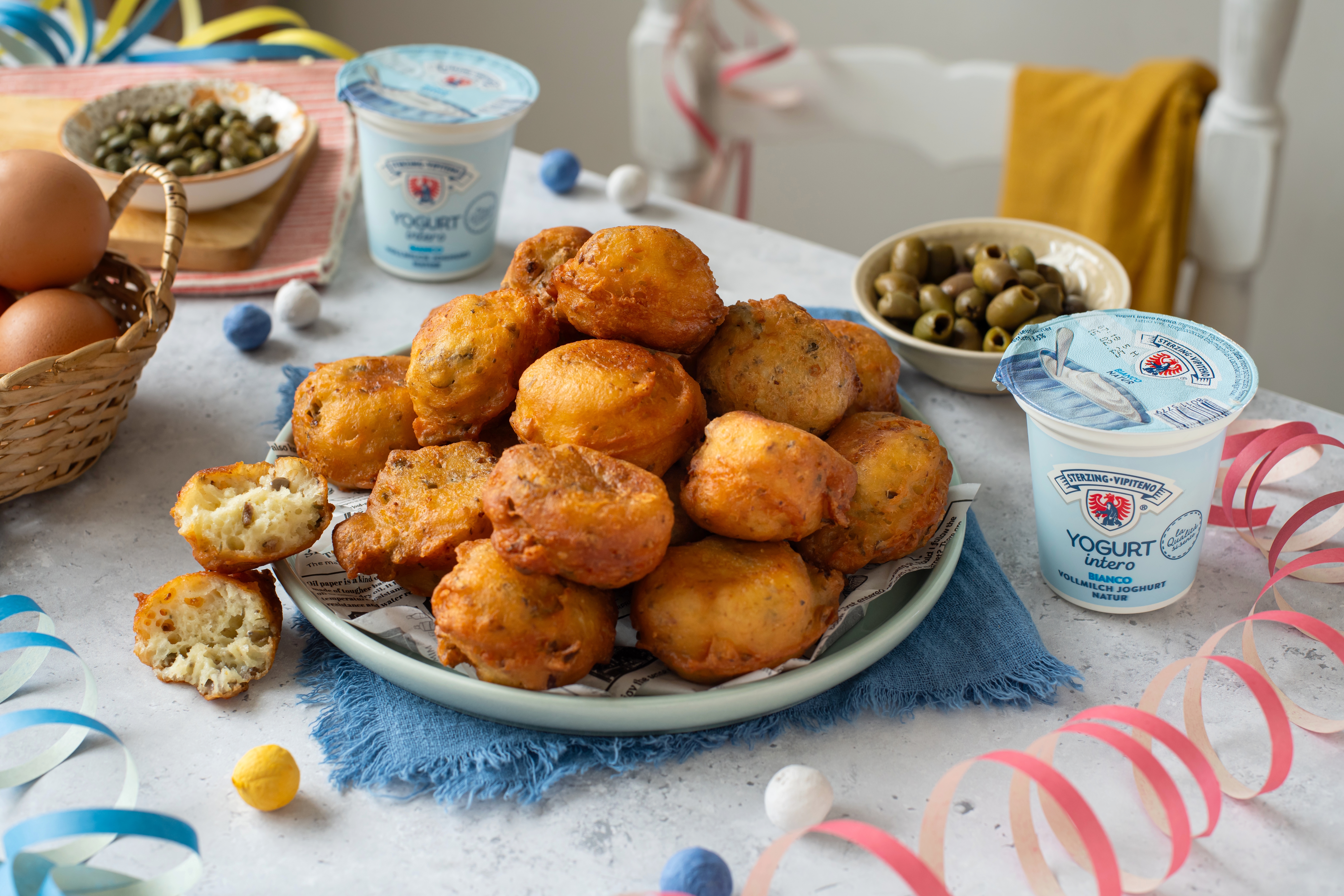 Teller mit frittierten Teigbällchen auf blauem Stoff, umgeben von Joghurtbechern, Oliven, Kapern, Eiern und bunten Papierstreifen