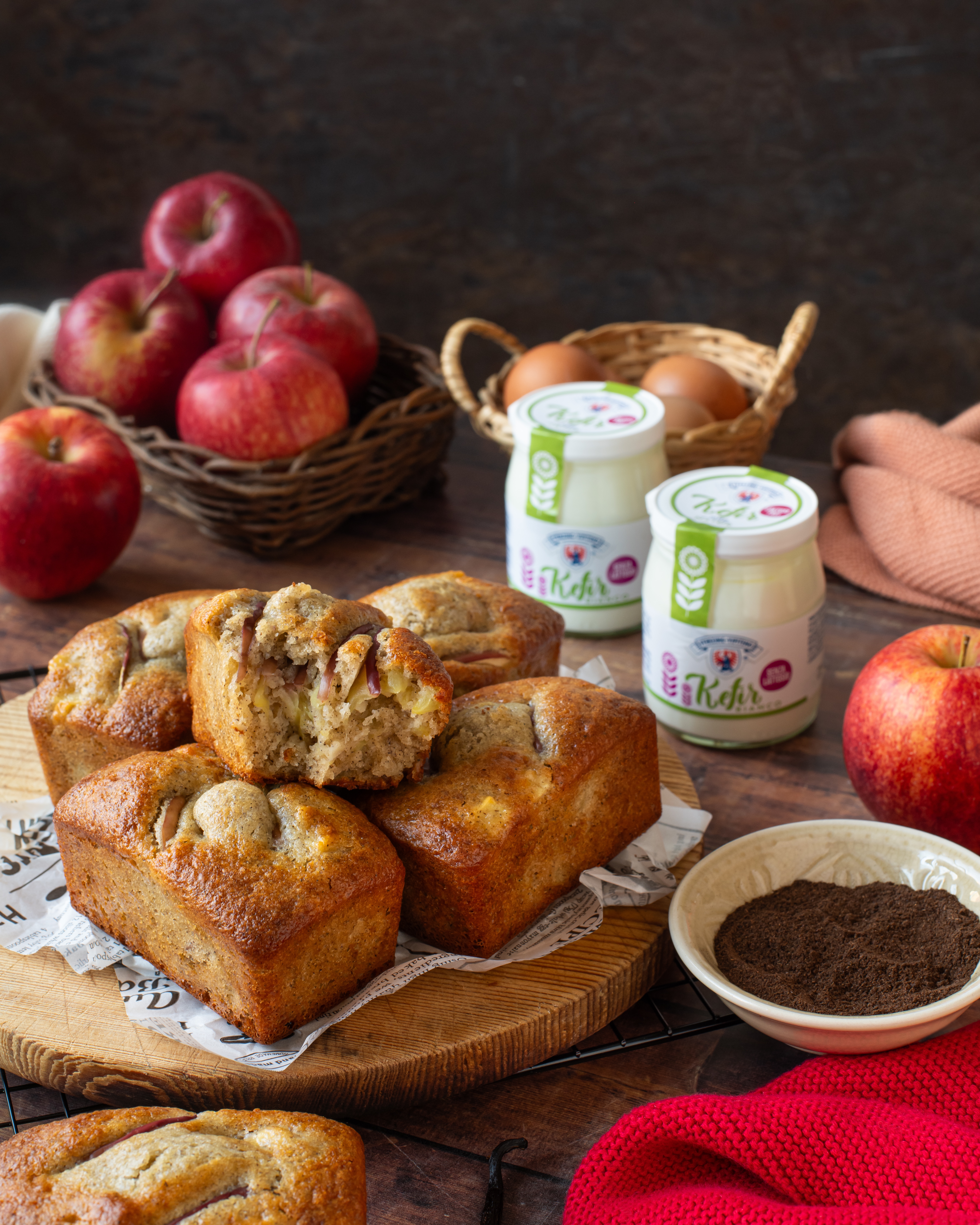 Mehrere kleine Kastenkuchen mit Apfelstücken auf einem Holzbrett neben Gläsern Kefir, Äpfeln, Eiern und einer Schale Vanillepulver