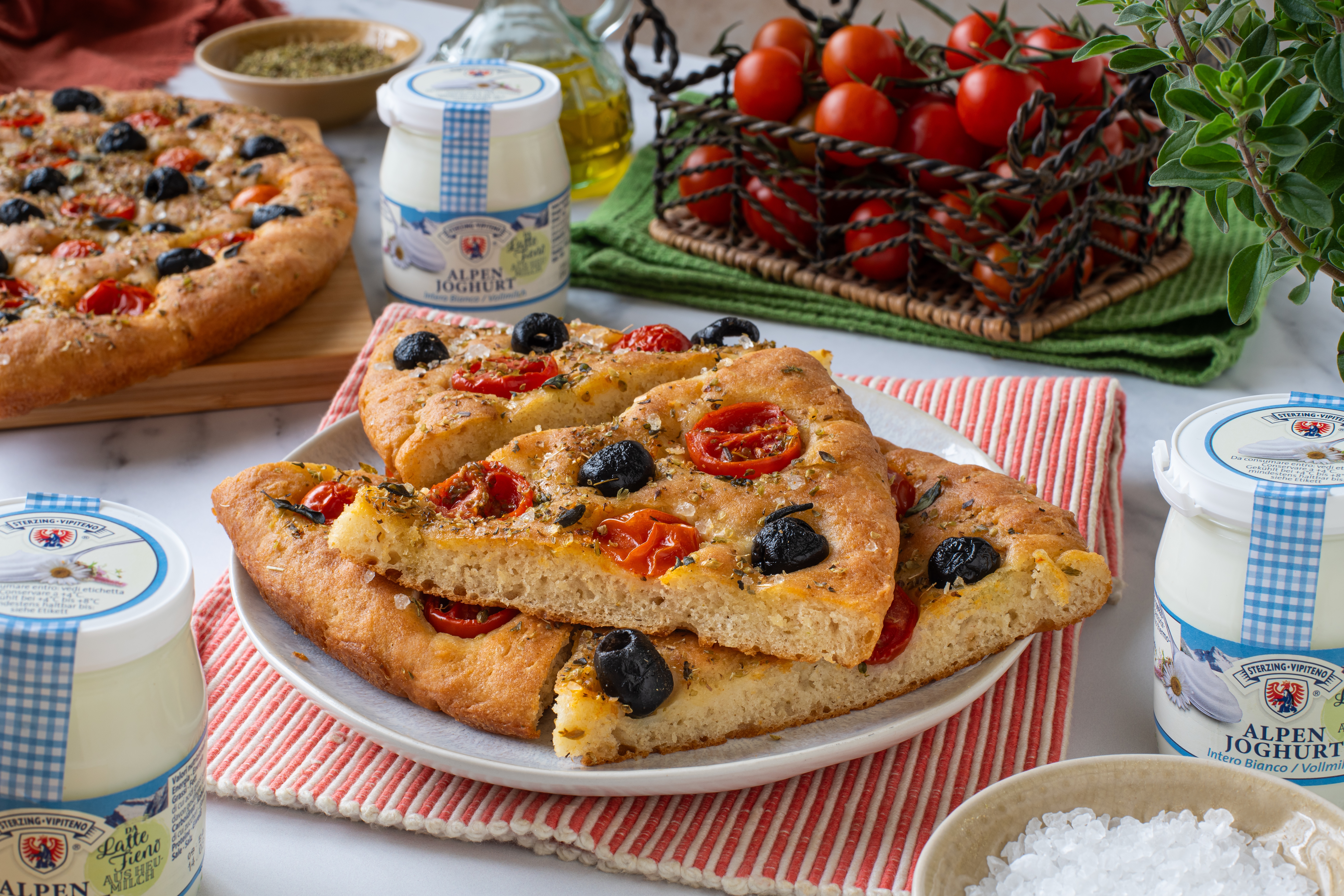 Dreieckige Stücke Focaccia mit Tomaten und schwarzen Oliven auf einem weißen Teller, umgeben von Joghurtgläsern und einem Korb mit Kirschtomaten